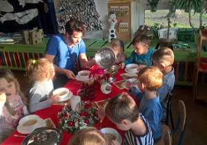 Na zdjęciu widać długi wigilijny stół, przy którym siedzą dzieci i wolontariusz, który próbuje naszych tradycyjnych potraw związanych ze Świętami Bożego Narodzenia.
